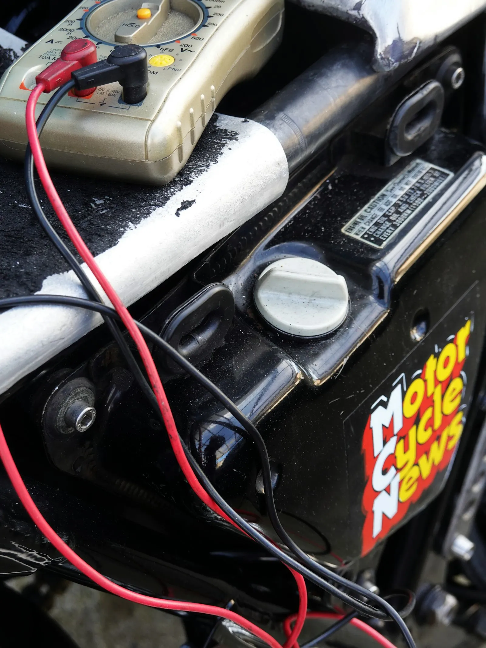 Multimètre posé sur une moto, ses câbles de mesure rouge et noir tendus le long du cadre et du cache latéral